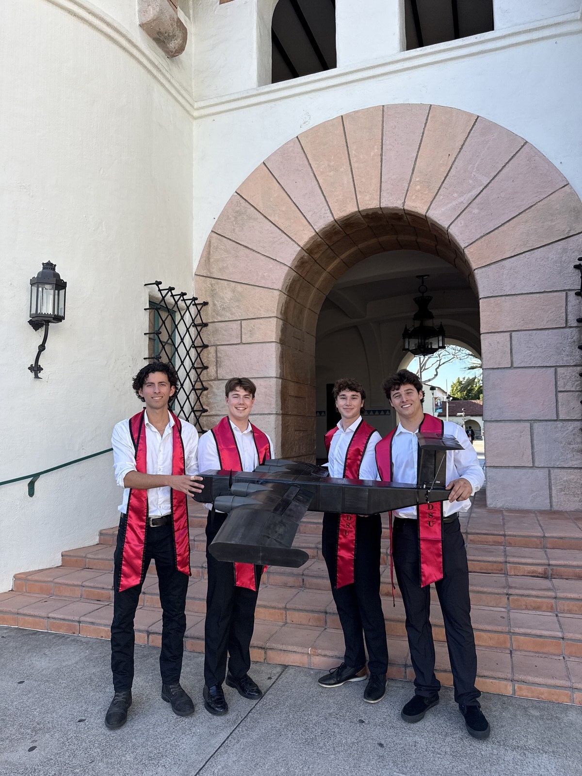 Team in graduation stoles holding the WIG vehicle in front of the SDSU campus arch.